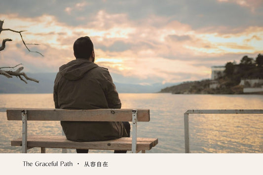 A person sitting alone on a bench, looking across a calm lake at sunset.