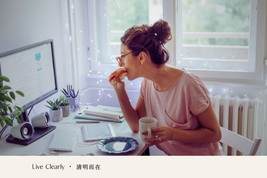 A person eating a sandwich while working at a desk with a computer and a cup of coffee.